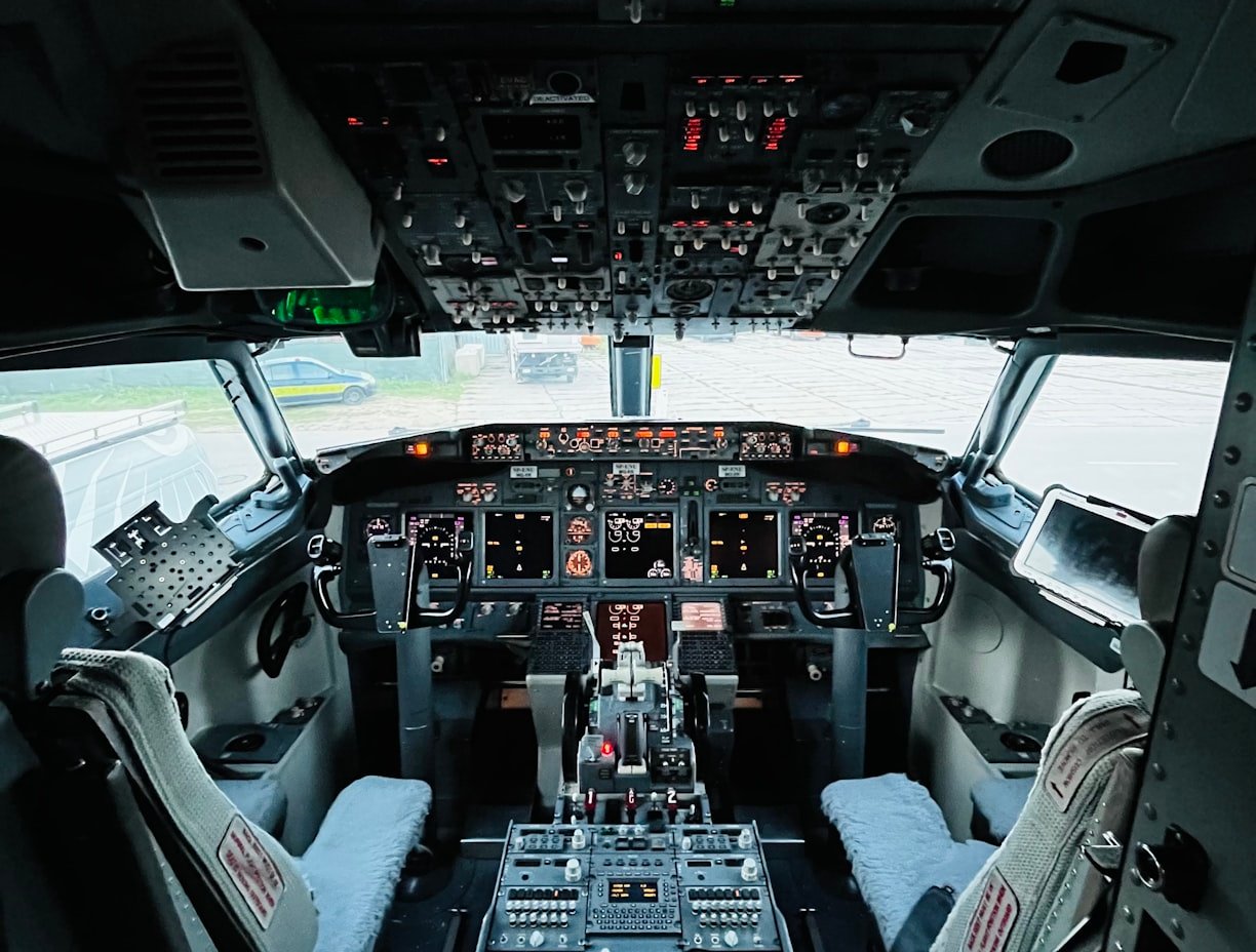 The cockpit of an aircraft with two pilot seats, a dashboard of screens and switches and a densely packed overhead panel.