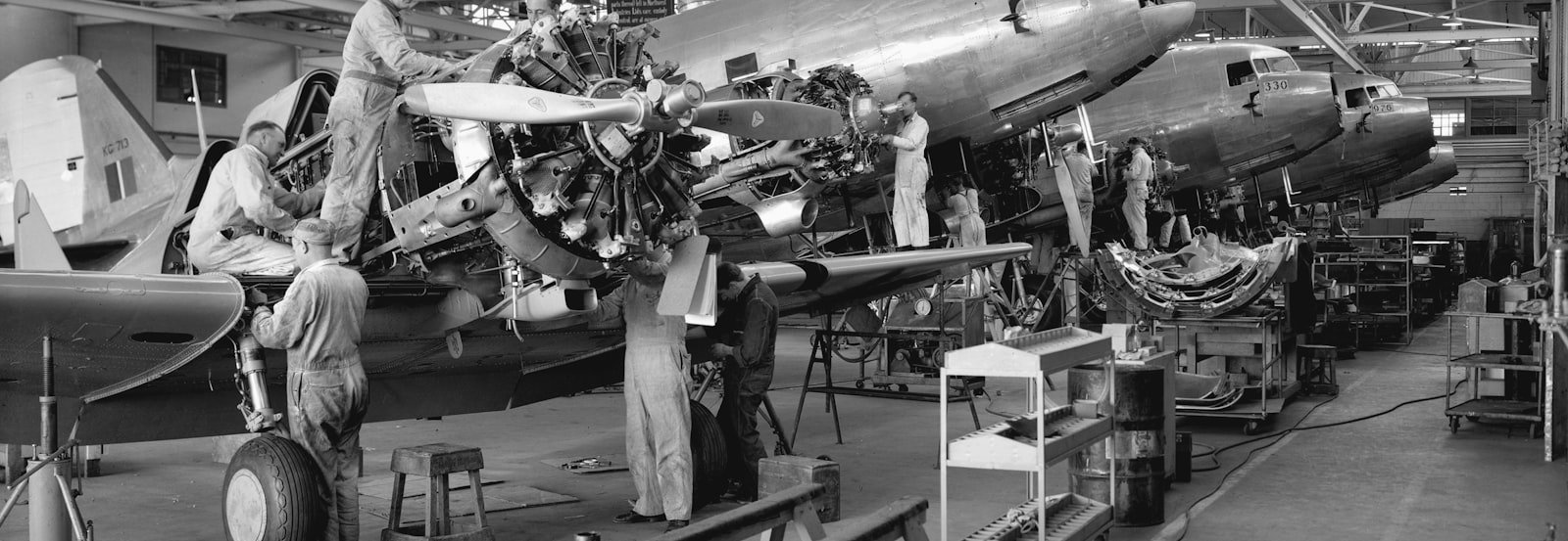 Workers maintaining and assembling aircraft inside an expansive hangar with large windows and overhead steel beams.