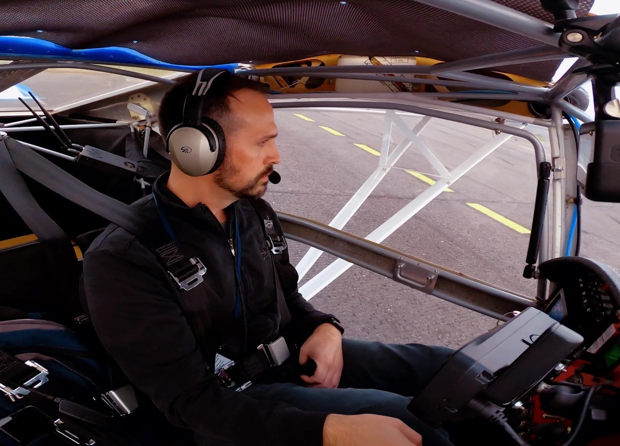 A person wearing a headset seated inside a small aircraft cockpit surrounded by controls, with a runway visible outside.