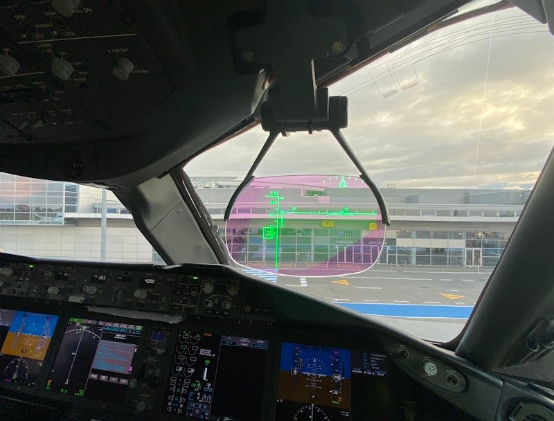 An aircraft cockpit with digital displays and a heads-up display showing flight information, with the airport terminal beyond.